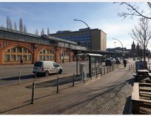 Straßenszene mit einem roten Backsteingebäude, das Eisenbahnarkaden enthält, und darüber verlaufenden Gleisen. Im Vordergrund sind ein Bürgersteig mit Fahrradständern, eine Bushaltestelle, parkende Autos und ein weißer Lieferwagen zu sehen, während im Hintergrund weitere Gebäude und Kirchtürme erkennbar sind.