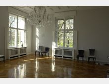 Heller Raum mit großen, weiß gerahmten Fenstern, einem polierten Holzboden, einem Kronleuchter an der Decke und mehreren Stühlen entlang der Wände. Durch die Fenster fällt Tageslicht, und draußen sind Bäume sichtbar.