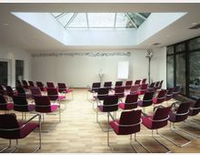Ein heller Konferenzraum mit Reihen aus roten Stühlen, einem Holzboden und einer großen Glasdachöffnung, die Tageslicht hereinlässt. Im Hintergrund befinden sich ein Rednerpult, ein Flipchart und schwarze Wanddekorationsmotive, rechts ist eine Fensterfront mit Blick ins Grüne.