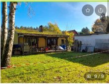 Ein kleiner gelber Bungalow mit einer überdachten Terrasse, umgeben von einem großzügigen Garten mit Bäumen und Rasen. Im Hintergrund sind Bäume und ein blauer Himmel sichtbar, rechts steht ein Nebengebäude.