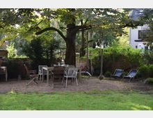 Grüner Garten mit einem großen Baum in der Mitte, darunter ein Holztisch mit verschiedenen Stühlen. Im Hintergrund sind Liegestühle, dichte Büsche, ein Holzzaun und ein Wohnhaus zu sehen.