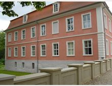 Zweistöckiges, rosafarbenes Gebäude mit symmetrisch angeordneten weißen Fensterrahmen und einem roten Ziegeldach. Im Vordergrund verläuft eine helle Steinmauer, und im Hintergrund sind Bäume zu sehen.