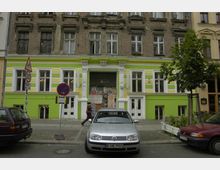 Fassade eines mehrstöckigen Altbaus mit unterem Bereich in auffälligen Grün- und Gelbtönen gestrichen, während der obere Teil aus unverkleidetem Backstein besteht. Vor dem Gebäude sind parkende Autos, Verkehrsschilder und ein einzelner Baum zu sehen.