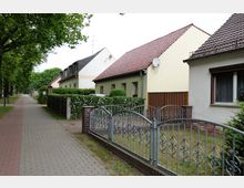 Eine ruhige Wohnstraße mit mehreren Einfamilienhäusern in heller Verputzoptik und roten Ziegeldächern. Die Häuser sind von gepflegten Hecken umgeben, und ein Bürgersteig verläuft parallel zu einer Baumreihe entlang der Straße.