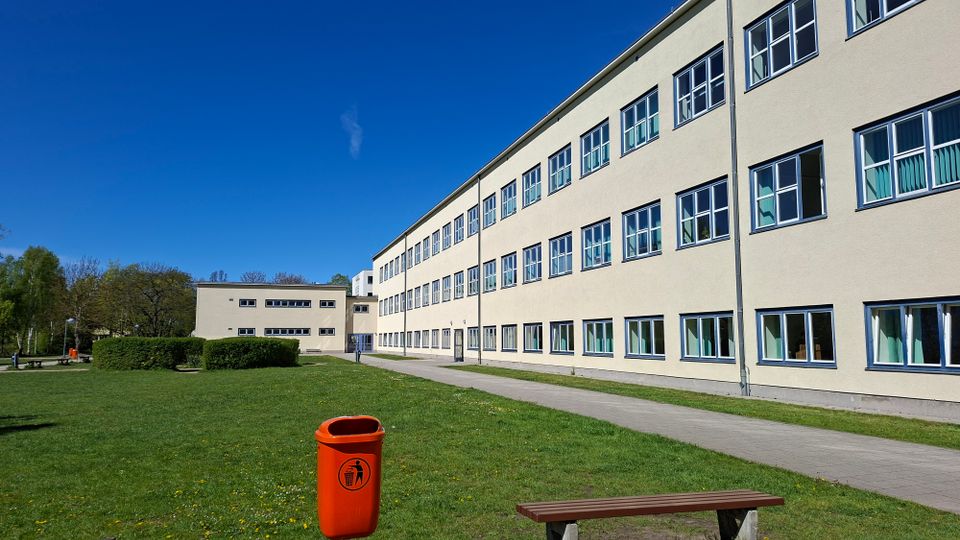 Großer, mehrstöckiger Gebäudekomplex mit heller Fassade und langen Reihen rechteckiger Fenster entlang eines Gehwegs. Davor liegen eine weitläufige Rasenfläche mit Hecken, einer Bank und einem orangefarbenen Abfallbehälter unter blauem Himmel.