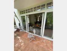 Terrasse mit roten Fliesen und weißen Metallstühlen, die zu einem Wohnraum mit großen Fenstertüren führt. Im Inneren sind ein Esstisch mit Stühlen, eine helle Deckenlampe und Regale sichtbar.