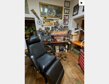 Innenraum eines Tattoo-Studios mit einem großen schwarzen Liegestuhl, Holzfußboden und Arbeitsflächen mit Lampen, Geräten und farbigen Tintenfläschchen in Regalen. Die Wände sind dicht mit gerahmten Bildern, Urkunden und Dekorationen versehen, neben Fenstern und einer Werkzeugkommode.