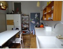 Kleine Küche mit Holzboden, einem weißen Esstisch mit Stühlen, einer Spüle mit weißer Arbeitsplatte und einer hölzernen Wandregaleinheit. Im Hintergrund sind ein Kühlschrank, ein Metallregal mit Büchern und Küchenutensilien sowie eine pinnwandartige Wand mit Magneten und Notizen sichtbar.