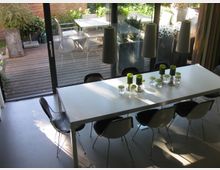 Esszimmer mit einem langen weißen Tisch und schwarzen Stühlen, dekoriert mit grünen Kerzen und Glasgefäßen. Im Hintergrund große Fensterfront mit Blick auf eine Holzterrasse und einen Garten mit Pflanzen.