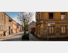 Straßenszene in einer historischen Wohngegend mit gepflastertem Weg, bunt gestrichenen Reihenhäusern auf der linken Seite und einer vergilbten, leicht verfallenen Hausfassade auf der rechten Seite. Ein kahler Baum und vereinzelte Laternen säumen die Straße, auf der ein weißer Transporter geparkt ist.