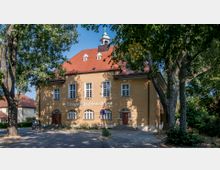 Historisches evangelisches Gemeindehaus mit gelber Putzfassade, roten Ziegeldach und kleinen Gauben, umgeben von hohen Bäumen und einer gepflasterten Straße im Vordergrund. Über dem Eingang ist der Schriftzug „Evangelisches Gemeindehaus“ sichtbar.