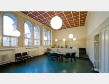 Innenraum eines historischen Gebäudes mit hohen Decken, großen Rundbogenfenstern und weißen Wänden mit Paneelverkleidung. Im Raum stehen ein runder Besprechungstisch mit Stühlen, daneben ein Empfangstresen und mehrere Hängelampen in Kugelform an der Decke.