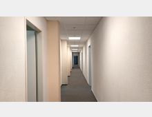 Langer, schlichter Flur in einem Bürogebäude mit weißen Wänden und hellem Teppichboden, Türen zu beiden Seiten und einer Kassettendecke mit eingelassenen Leuchten. Am Ende des Flurs befindet sich eine geschlossene Glastür.