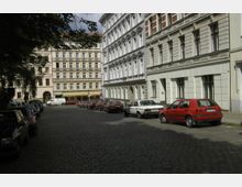 Straßenansicht mit gepflasterter Fahrbahn und geparkten Autos an beiden Seiten, umgeben von mehrstöckigen Wohnhäusern mit verzierten Fassaden und großen Fenstern. Im Hintergrund sind weitere Gebäude und ein Baum erkennbar.