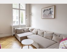 Wohnzimmer mit hellbeiger Eckcouch und kleinen Kissen vor einer großen weißen Doppelfensterfront. An der Wand hängt ein gerahmtes Poster, und der Holzboden ist in Fischgrätmuster verlegt.