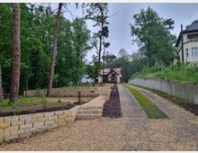 Ein geschotterter Weg führt durch eine bewaldete Umgebung zu einem modernen Holzhaus mit roten Dachziegeln. Links des Weges befinden sich Terrassenmauern aus Naturstein, während rechts ein Hang mit Betonmauer und Grasbepflanzung verläuft.