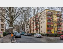 Wohnstraße mit mehrstöckigen Wohngebäuden auf beiden Seiten, die gelb und beige gestrichen sind und rot-orangene Balkone haben. Entlang der Straße stehen Bäume ohne Laub, geparkte Autos sowie ein Gehweg mit Straßenlaternen und einem Straßenschild mit der Aufschrift „Landshuter Straße“.