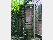 Grüne Wendeltreppe aus Metall mit goldenen Details, die an einer rosa Holzfassade eines Gebäudes angebracht ist, umgeben von Efeu und Pflanzen im Außenbereich.