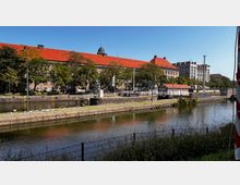 Historisches Gebäude mit rotem Ziegeldach und einer Kuppel im Hintergrund, umgeben von Bäumen entlang eines Wasserkanals mit Schleusenanlage im Vordergrund. Der Kanal ist von Grünflächen und einem Zaun gesäumt, und moderne Gebäude sind im rechten Bildbereich sichtbar.