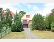 Einfamilienhaus mit Satteldach und weißer Fassade, einer Garage im Erdgeschoss sowie geschlossenen Rollläden; umgeben von einem Garten mit Bäumen und Sträuchern, einer gepflasterten Einfahrt und einem Zaun zur linken Seite.