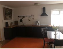 Eine moderne Küche mit dunklen Schränken, einer Arbeitsplatte aus Holz, einer Dunstabzugshaube und weißen Fliesen an der Wand. Im Vordergrund steht ein Esstisch mit einer Vase voller Tulpen, beleuchtet durch ein Fenster auf der rechten Seite, das natürliches Licht hereinlässt.