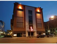 Backstein-Gebäude mit großen Gitterelementen und beleuchteter Glasfront, bei Dämmerung fotografiert. Vor der Fassade befindet sich ein leuchtendes rotes Schild mit der Aufschrift „Büro“, darunter sind parkende Autos und eine Straße sichtbar.