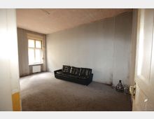 Ein spärlich eingerichteter, einfacher Raum mit kahlen, grauen Wänden und einem Teppichboden. Es gibt ein großes Fenster mit Heizung darunter, eine schwarze Ledercouch und eine Stehlampe mit gemustertem Lampenschirm in der Ecke.