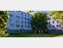 Mehrstöckiges Wohngebäude mit hellblauer Fassade und rechteckigen Fenstern, umgeben von einer Grünfläche mit Bäumen. Im Vordergrund befindet sich eine Wäscheleine aus Metallgestellen.