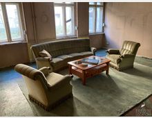 Karger, altmodisch eingerichteter Wohnraum mit großem Sofa und zwei Sesseln um einen Holz-Couchtisch auf einem grünen Teppich. Hohe Fenster lassen Tageslicht in den sonst spärlich möblierten Raum mit abgenutzten Wänden und Boden.