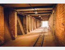 Ein unterirdischer, gemauerter Tunnel mit Backsteinwänden und sichtbaren Holzbalken an der Decke. In der Mitte verläuft ein Schienenstrang, der in ein hell erleuchtetes Ende des Tunnels führt.