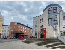 Straßenszene mit moderner Architektur: Im Vordergrund eine breite Treppe, rechts ein helles Gebäude mit großen, rot umrahmten Fenstern und einer Sparkassenfiliale im Erdgeschoss. Links ein dreistöckiges, terrakottafarbenes Gebäude mit der Beschriftung „Amtsgericht“, daneben ein grau verputztes Haus, vor dem Autos und ein roter Bus parken.