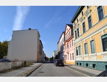 Straßenansicht mit mehrstöckigen Altbauten, rechts in gelb und rosa gestrichen, teilweise mit dekorativen Fassadenelementen. Links steht ein schmuckloses, weißes Gebäude; auf beiden Straßenseiten geparkte Autos und Gehwege, im Hintergrund teils Bäume sichtbar.