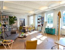 Heller, offener Raum mit weißen Backsteinwänden und Holzboden, eingerichtet mit einem grauen Sofa, einem runden Holztisch, farbigen Stühlen, Regalen voller Bücher und einem türkisfarbenen Schrank. Großer Lichteinfall durch mehrere Fenster, ergänzt von Pflanzen und einer Flipchart-Tafel.