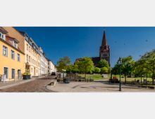 Straßenszene mit Kopfsteinpflaster, links eine Reihe von mehrstöckigen, farbigen Wohnhäusern und rechts eine Grünfläche mit Bäumen und Sitzbänken, im Hintergrund eine Backsteinkirche mit hohem Turm und spitzer Spitze. Der Himmel ist klar und blau.