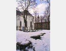 Schneebedeckter Friedhof mit Grabsteinen und Kreuzen vor einer weißen Kirche mit hohen Fenstern; im Hintergrund sind weitere Gebäude sowie ein Fernsehturm sichtbar, umgeben von unbelaubten Bäumen.