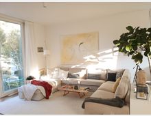 Helles Wohnzimmer mit einem großen, beigefarbenen Ecksofa, dekoriert mit Kissen in geometrischen Mustern. Im Raum befinden sich ein Couchtisch aus Holz, eine Zimmerpflanze, ein großes abstraktes Gemälde an der weißen Wand und eine Glastür, die auf einen Außenbereich führt.