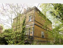 Gelbes Backsteingebäude in einem überwucherten Garten mit Bäumen und Kletterpflanzen, das architektonische Details wie verzierte Fensterrahmen und eine Balustrade aufweist. Das Gebäude wirkt von Vegetation teilweise verdeckt und ist von Tageslicht durchdrungen.