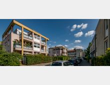 Mehrfamilienhäuser in moderner Bauweise mit breiten Balkonen, umgeben von Hecken und einer schmalen Straße, an der Autos geparkt sind; der Himmel ist klar mit vereinzelten Wolken.