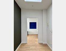 Flur eines modernen Raums mit hellem Holzfußboden, weißen Wänden und einem olivgrünen Akzent an der linken Wand. Im Hintergrund ist ein weiterer Raum mit einem großen blauen Bild an der Wand zu sehen.