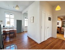 Helle Wohnung mit Holzdielenboden, weißen Wänden und hohen Decken. Im Vordergrund ein Essbereich mit Tisch und Stühlen, daneben ein grauer Schrank vor einem großen Fenster; rechts ein Flur mit weißen Türen, daneben ein Einblick in einen Nebenraum mit Regalen.