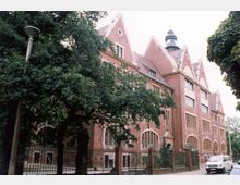 Historisches Backsteingebäude mit steilen Giebeln und spitzem Turm. Das Gebäude ist von Bäumen umgeben, mit einem Metallzaun im Vordergrund und einer Straße mit parkenden Fahrzeugen.