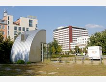 Flache städtische Parkfläche mit einem kleinen, geschwungenen Gebäude aus Metall und Glas im Vordergrund. Im Hintergrund stehen mehrstöckige Büro- und Wohngebäude, darunter ein großes, rot-weißes Hochhaus.