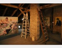 Ein rustikaler Dachboden mit Holzbalken und Holzfußboden, auf dem mehrere Gemälde ausgestellt sind. Zwei Leitern lehnen an einem Backsteinpfeiler; die ausgestellten Kunstwerke zeigen figurative und abstrakte Motive.