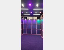 Holzrahmenstruktur mit dunklen, blauen Wänden, beleuchtet durch farbige LED-Lichter in Lila, Rot, Blau und Grün an der Decke. Der Boden ist mit einem dunkel grauen Teppich bedeckt, und oben an der Rückwand sind Schilder mit der Aufschrift "B1" angebracht.