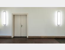 Eine weiße Doppeltür in einer schlichten Wand, flankiert von zwei vertikalen Wandleuchten mit hellem Licht. Der Boden besteht aus dunklen, glänzenden Fliesen mit einem niedrigen Sockel aus hellem Stein.