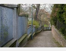 Gepflasterter Gehweg mit leichtem Gefälle, eingefasst von einer grauen Betonsäulen- und Metallzaunkonstruktion auf der linken Seite und einer Backsteinmauer mit darüberliegendem Maschendrahtzaun auf der rechten Seite; umgeben von Bäumen und Sträuchern. Im Hintergrund sind Dächer von Gebäuden sichtbar.