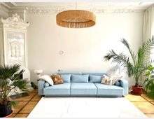 Helles Wohnzimmer mit einer pastellblauen Couch, dekorativen Kissen und einem verzierten weißen Kachelofen mit Relief. Der Raum ist mit Zimmerpflanzen, einer geflochtenen Deckenlampe und einem hellen Vorhang gestaltet.