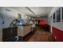 Ein schlichter Innenraum mit Holzboden und einer Bar an der linken Seite, dekoriert mit hängenden Lampen. Runde Bögen in der Wand bieten Durchblicke, und an der roten Rückwand befinden sich Sitzmöglichkeiten; Poster hängen an der weißen und roten Wand rechts.