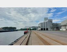 Industriehafen mit breitem Kai und Schienen auf der Pier, rechts ein großes graues Industriegebäude mit Förderanlagen und Lagerhallen. Links ein Fluss mit einem Anlegesteg und angrenzenden Bäumen im Hintergrund unter teils bewölktem Himmel.