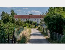 Schmaler Weg, flankiert von dichtem Grün und niedrigen Gartenzäunen, führt zu einer Reihenhausanlage mit roten Ziegeldächern und weißen Fassaden im Hintergrund. Der Himmel ist leicht bewölkt, und zwei Vögel fliegen über die Szene.